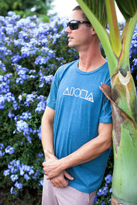 Man leaning against a tropical palm tree wearing our Mauka to Makai Aloha Shapes sueded jersey tee in heather deep teal