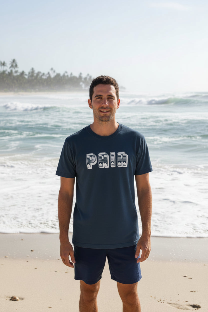A man standing in front of a beach with waves in background, wearing a blue t-shirt with 'PAIA' printed on the front and blue shorts.