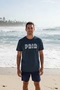 A man standing in front of a beach with waves in background, wearing a blue t-shirt with 'PAIA' printed on the front and blue shorts.