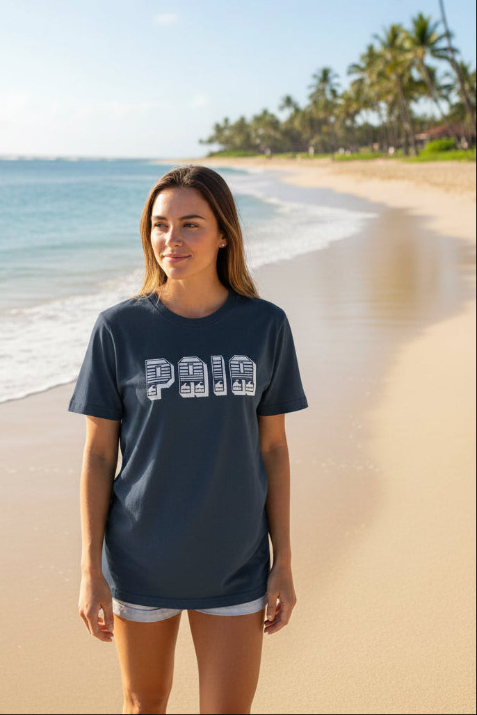 A woman stands on a sandy beach wearing a vintage denim blue t-shirt with a white graphic print that reads 'PAIA'. The background features a tropical beach scene with palm trees, blue ocean water, and a clear sky.