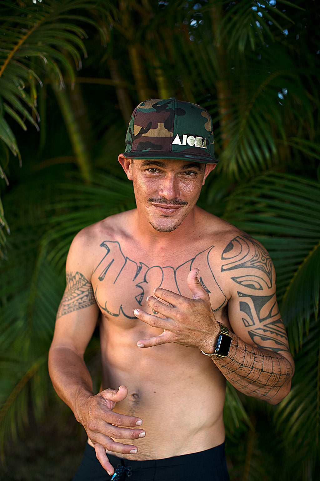 Man with tattoos wearing a camouflage print Aloha Shapes ® snapback hat in front of palm trees