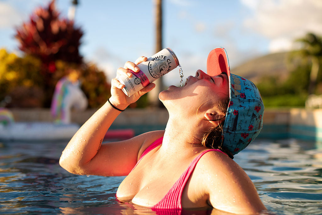 Young lady wearing Aloha Shapes ® snapback hat in a pool drinking a White Claw beverage