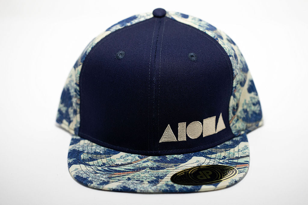 Adult flat brim snapback hat. Navy blue front panel embroidered with white ALOHA Shapes ® logo. Brim and back panels are printed with Japanese waves from old woodblock paintings.
