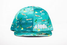 Adult flatbrim snapback hat with blue satin fabric printed with airplanes, aloha, Pacific and palm trees all over. Embroidered with white Aloha Shapes logo