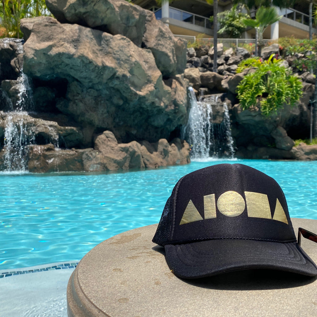 Adult Aloha Shapes ® black trucker hat foil printed with metallic gold logo on a table in front of a tropical pool in Hawaii