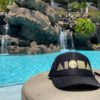 Adult Aloha Shapes ® black trucker hat foil printed with metallic gold logo on a table in front of a tropical pool in Hawaii