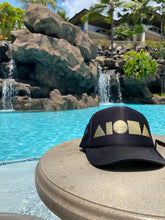 Adult Aloha Shapes ® black trucker hat foil printed with metallic gold logo on a table in front of a tropical pool in Hawaii