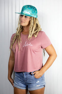 Woman with blond dreadlocks wears Adult flatbrim snapback hat with blue satin fabric printed with airplanes, aloha, Pacific and palm trees all over. Embroidered with white Aloha Shapes logo