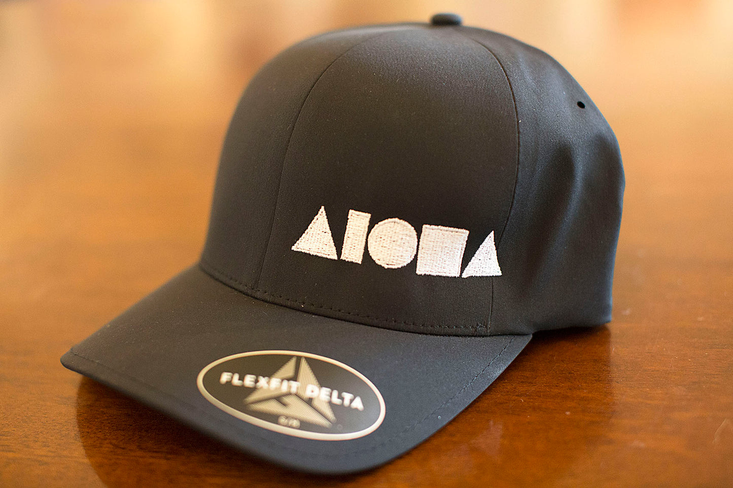 Black ALOHA Shapes ® logo Flexfit hat sitting on a wooden desk