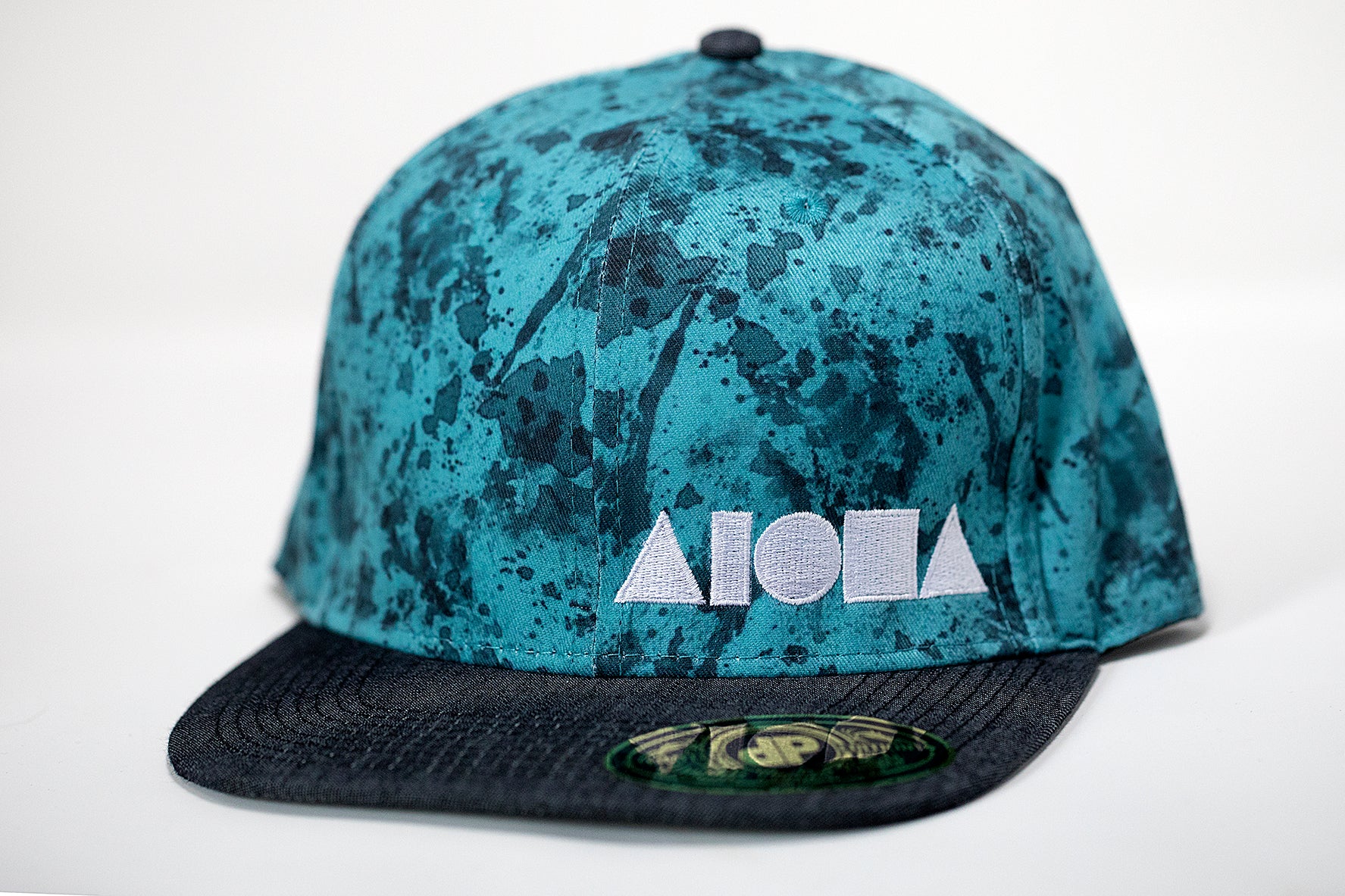 Adult flat brim snapback hat. Grey denim brim. Blue fabric with a paint splatter-like print. Embroidered with white ALOHA Shapes ® logo