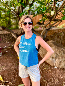 Woman smiling in front or a rock wall wearing Humble Kind Aloha womens crop tank top.