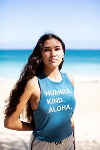 Woman on tropical beach in front of blue water wearing blue Humble Kind Aloha crop racerback tanktop