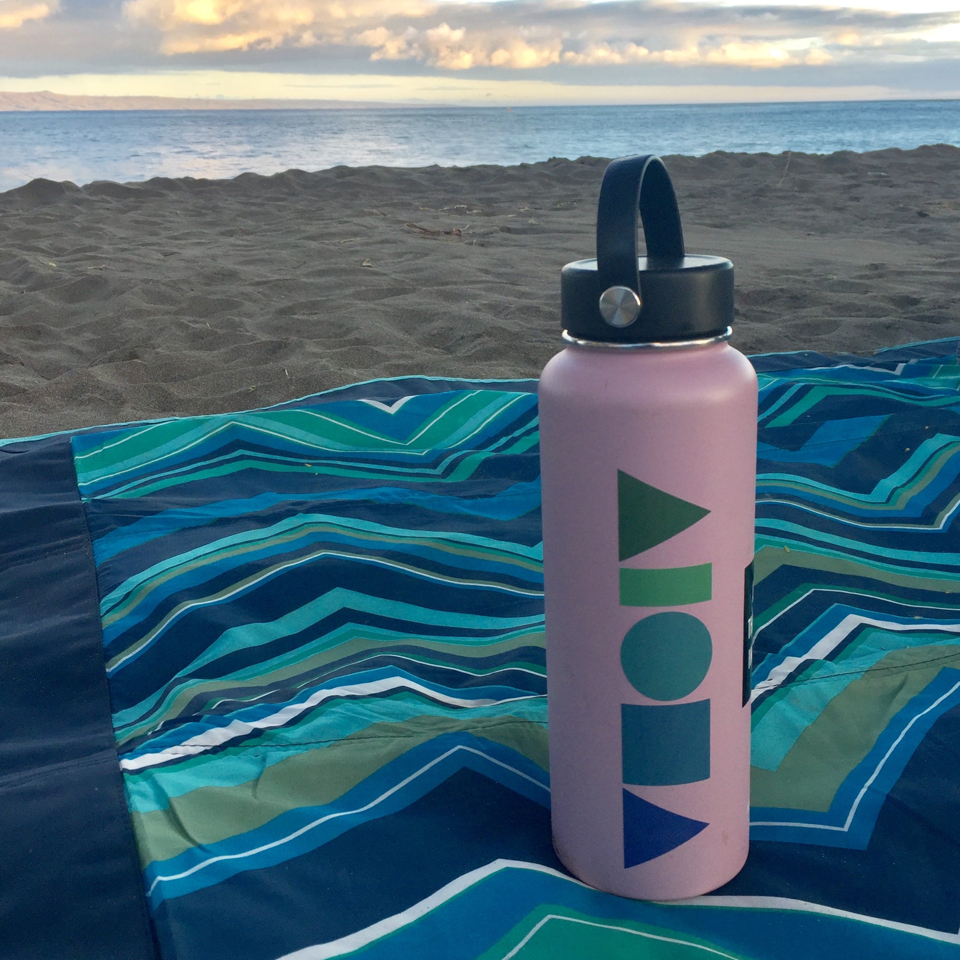 Hydroflask water bottle with a Blue Dream Aloha Shapes logo sticker on a beach blanket with Hawaii sunset in background