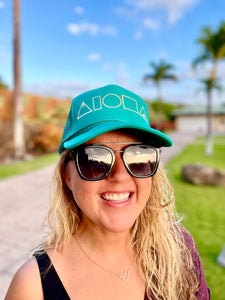 Woman wearing sunglasses and Mauka to Makai Aloha Shapes® adult trucker hat with tropical palm trees in background