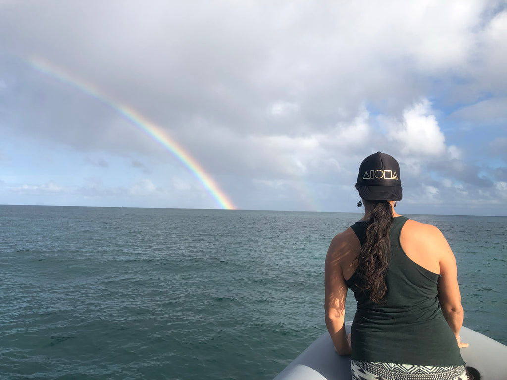 Person on an inflatable raft watching a rainbow with Mauka to Makai Aloha Shapes trucker hat turned backwards