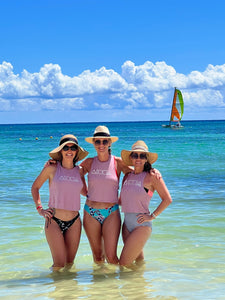 Three ladies on a tropical beach wearing Mauka to Makai Aloha Shapes womens crop racerback tanks with sailboat in the background