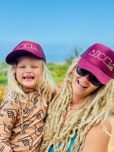 Mother and daughter wearing matching maroon and gold trucker hats foil printed with Mauka to Makai Aloha Shapes logo in metallic gold