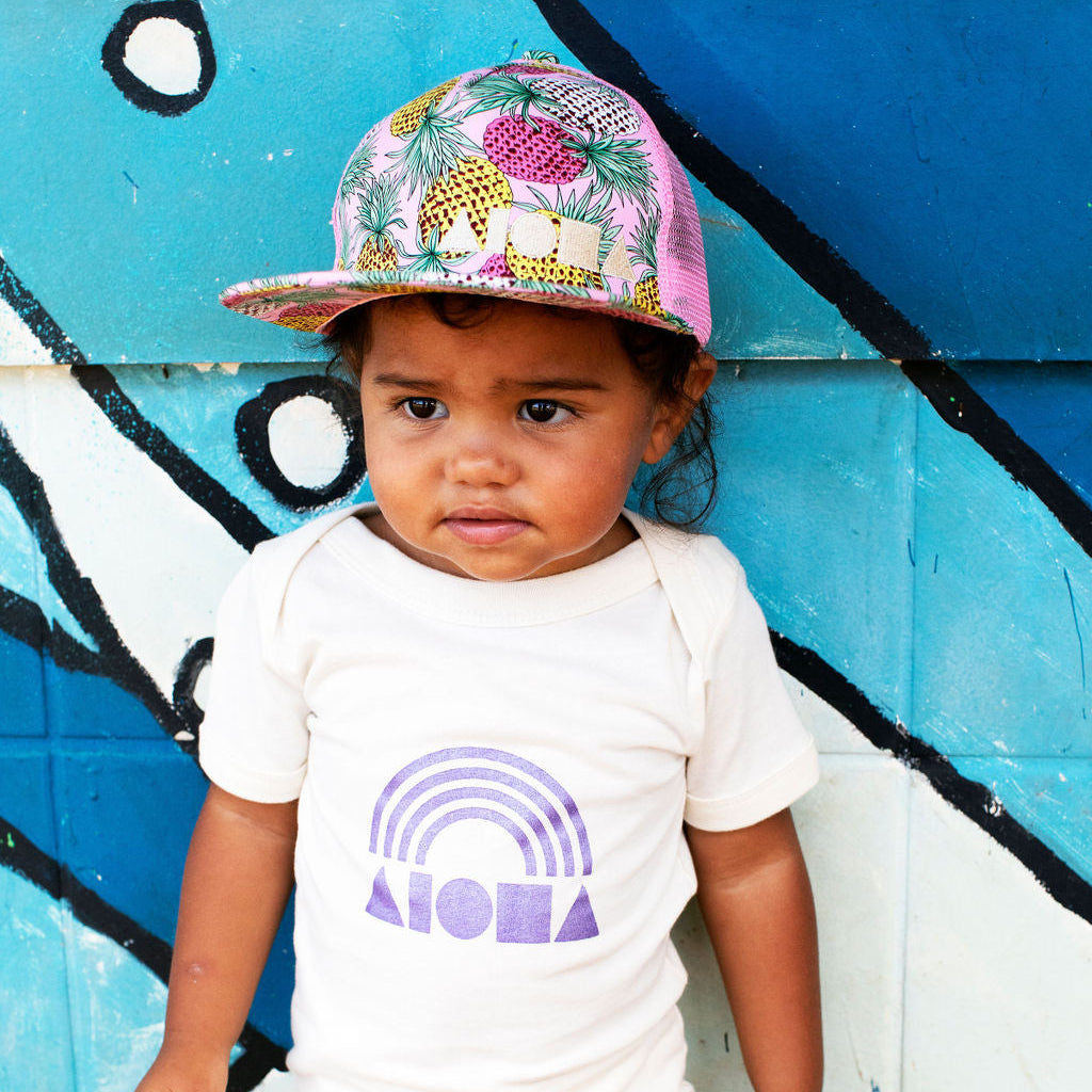 Baby girl wearing a Pink Pineapple Aloha Shapes hat in front of a graffiti wall
