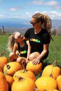 Mom and daughter in pumpkin patch Upcountry Maui. Mom is wearing black unisex tee screen printed with rainbow color Aloha Shapes® logo
