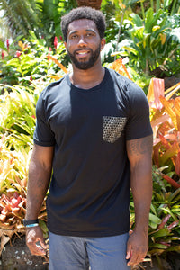 Man wearing black pocket tee printed with Aloha shapes logo repeated standing in front of tropical foliage
