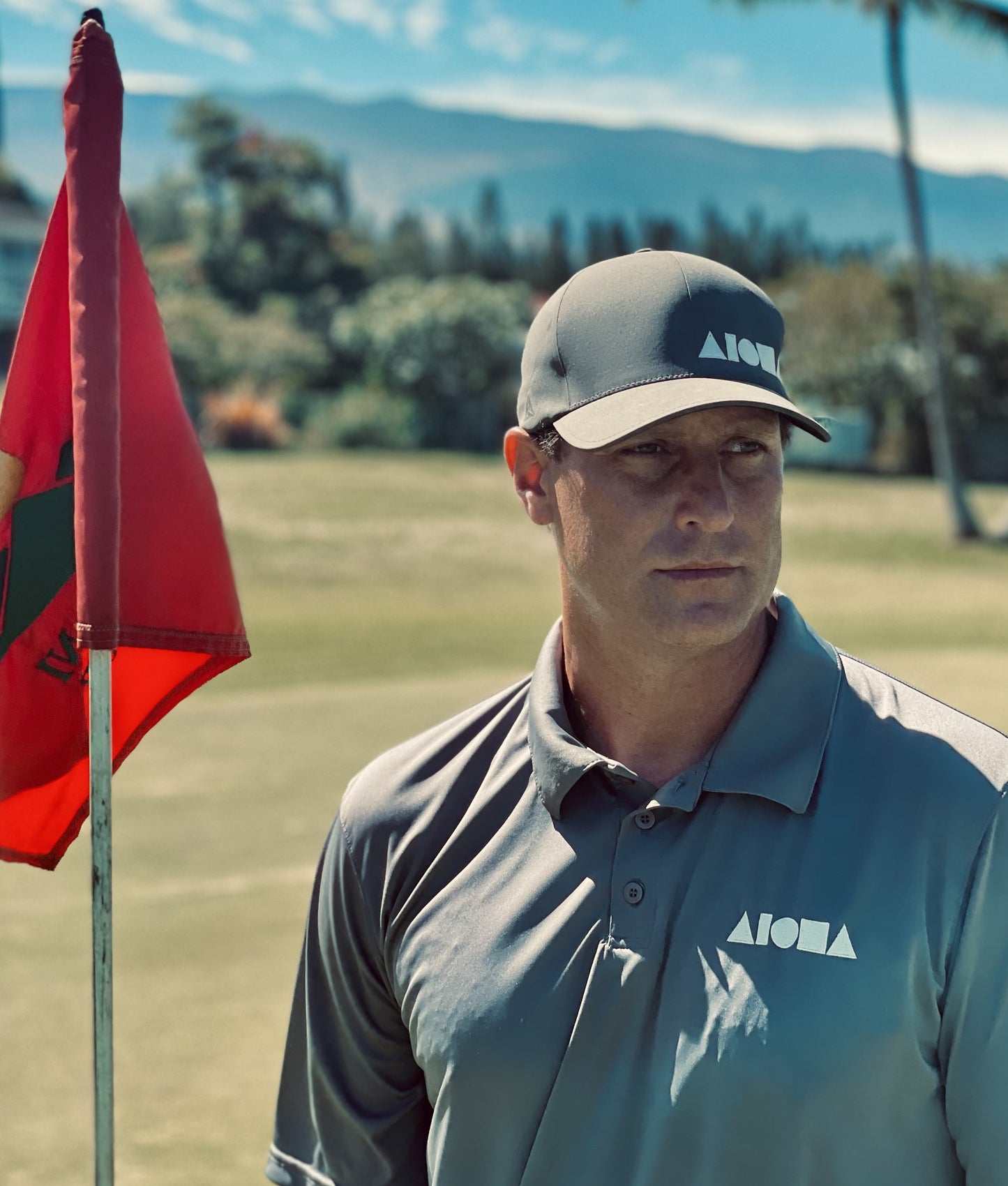 Person standing by a golf flag on the green wearing an embroidered Aloha Shapes® collared polo shirt and a Light grey Flexfit hat with white Aloha Shapes® logo