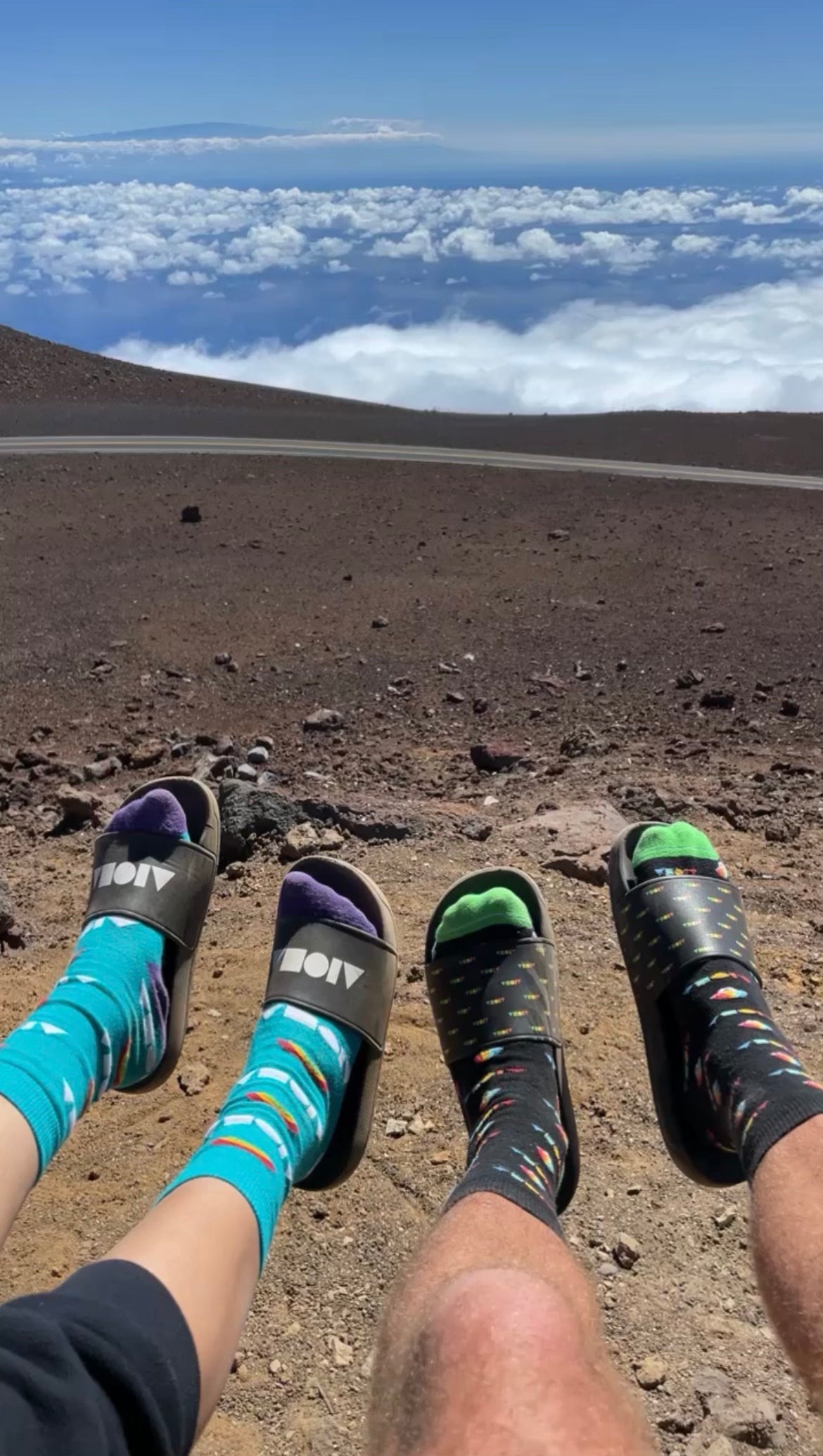 Two pairs of feet wearing Aloha Shapes® slide slippahs hanging over the edge of scenic lookout at Haleakala Maui hawaii
