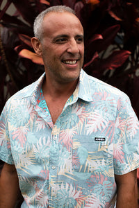 Man standing in front of tropical plants smiling wearing a "Backyards" aloha surf shapes collared aloha shirt