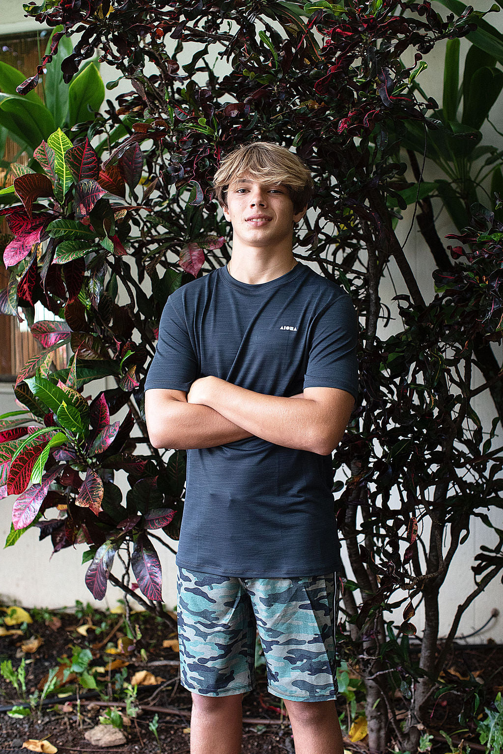 Surfer boy wearing camo print unisex board shorts in front of tropical foliage in Hawaii