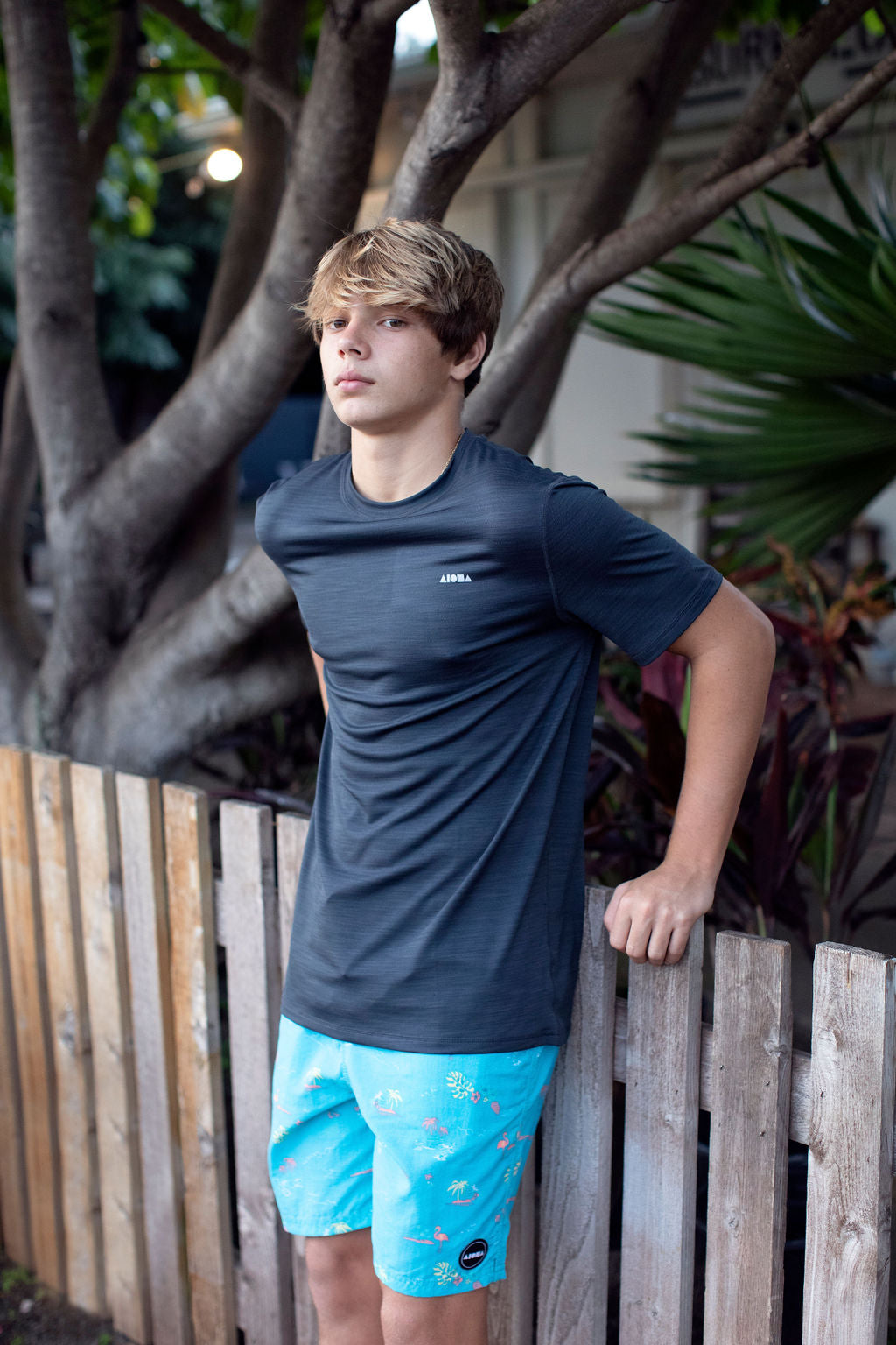 Young surfer leaning against wooden fence wearing Aloha Surf Shapes "Shorey" youth swim trunks