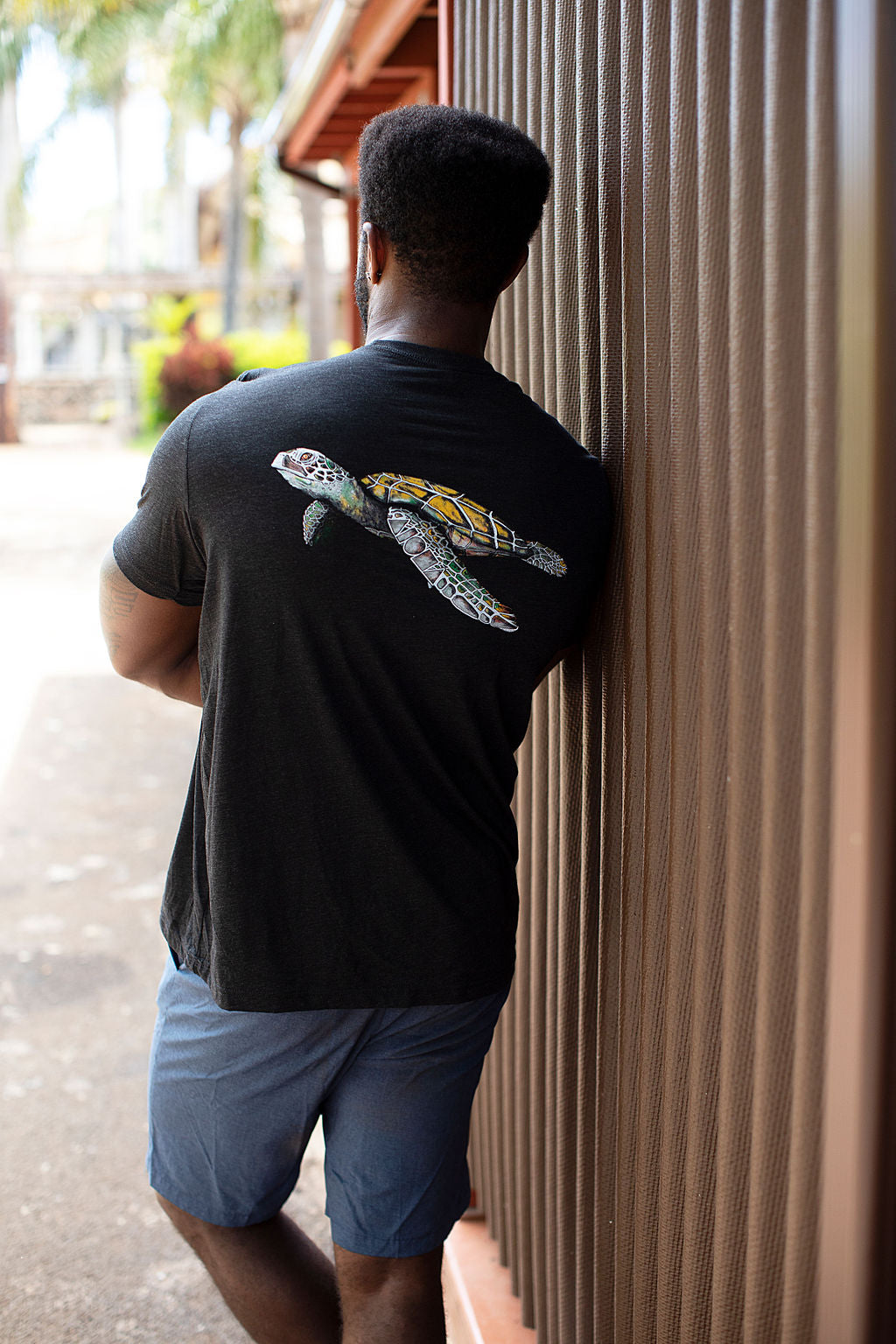 Tall man wearing an Aloha Shapes ® Turtle design t-shirt leaning against a wall with his back turned to show the tshirt