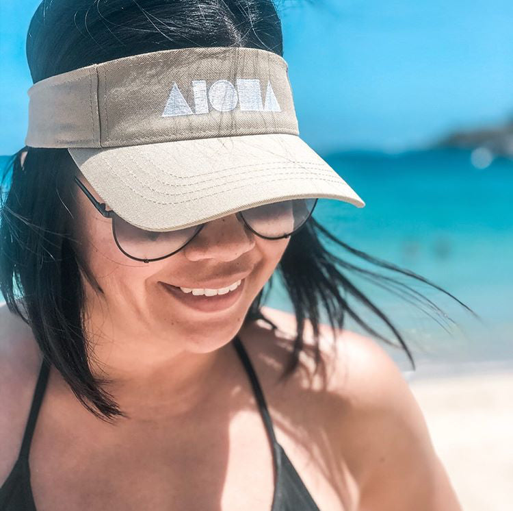 Woman with ocean in background wearing a khaki color Aloha Shapes visor. Logo is white