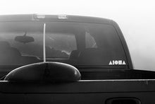 Black and white photo of a pickup truck with surfboard hanging out the back. White ALOHA Shapes ® logo decal sticker on back window of truck. 