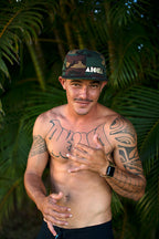Man with tattoos wearing a camouflage print Aloha Shapes ® snapback hat in front of palm trees